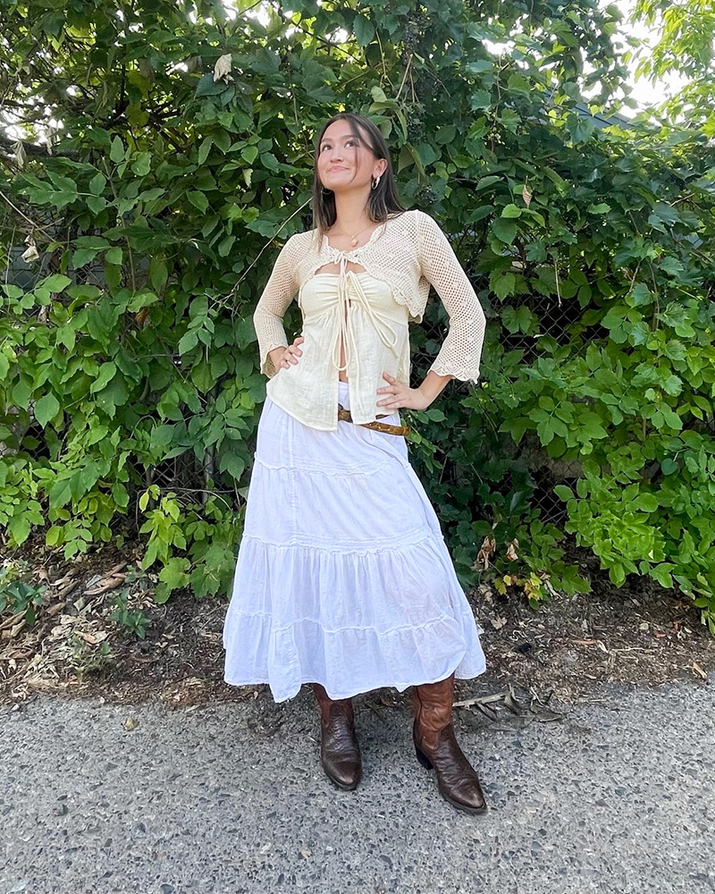 Woman wearing cream sweater and white maxi skirt stands in front of green bush