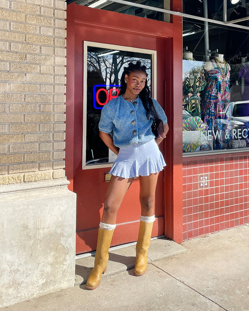 A woman wearing a cropped blue sweater, blue shorts and tan Frye Campus boots