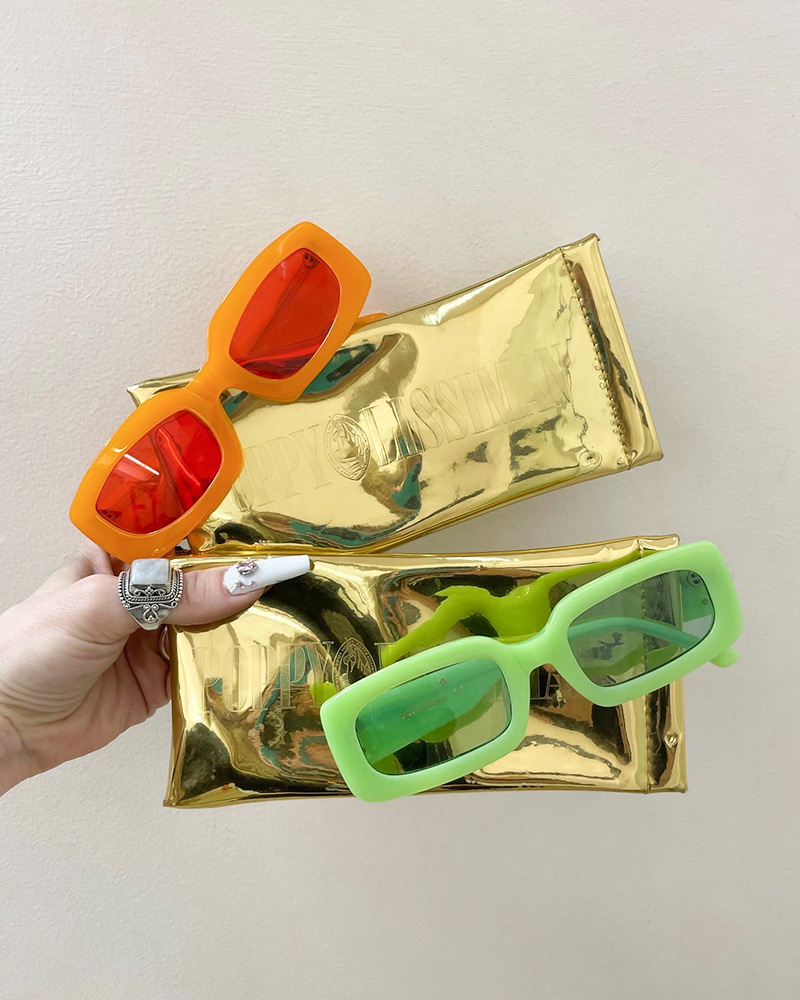 A hand holds two pairs of Poppy Lissman orange and green sunglasses and their gold foil carrying cases