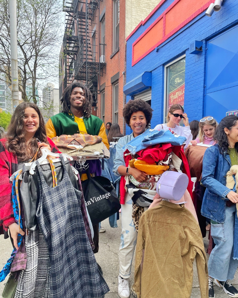 three friends smile while holding armfulls of clothing near a blue brick building