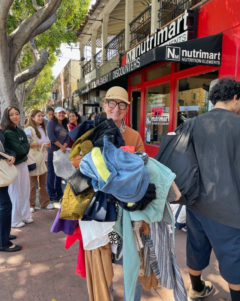 person standing in front of line of shoppers on sidewalk holds pile of clothes